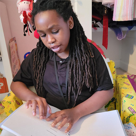 Photo of 12 year old girls sitting on a yellow chair reading a braille book.