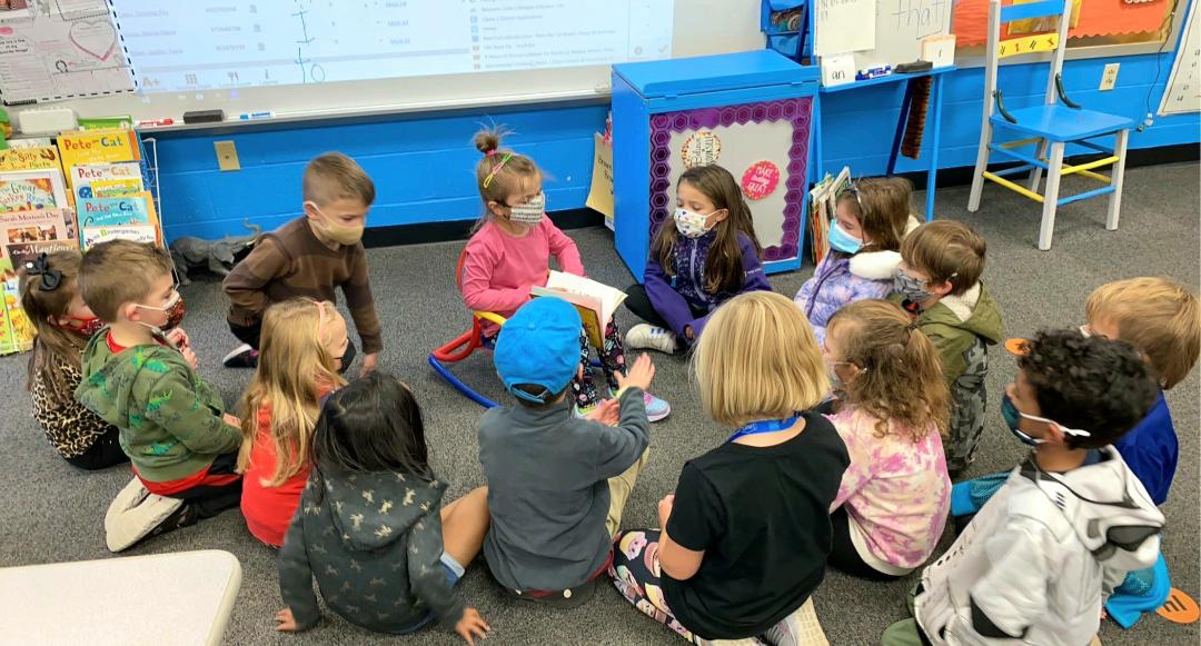 DEI-photo group of children in a circle around a young girl reading a braille book.