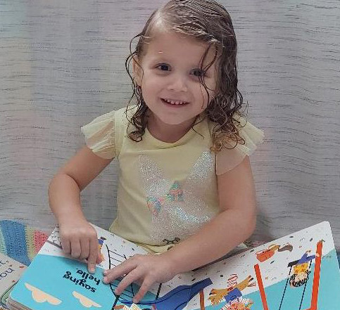 An enthusiastic young girl with long brown hair, wearing a colorful dress, joyfully holds a Braille book in a library setting filled with books.