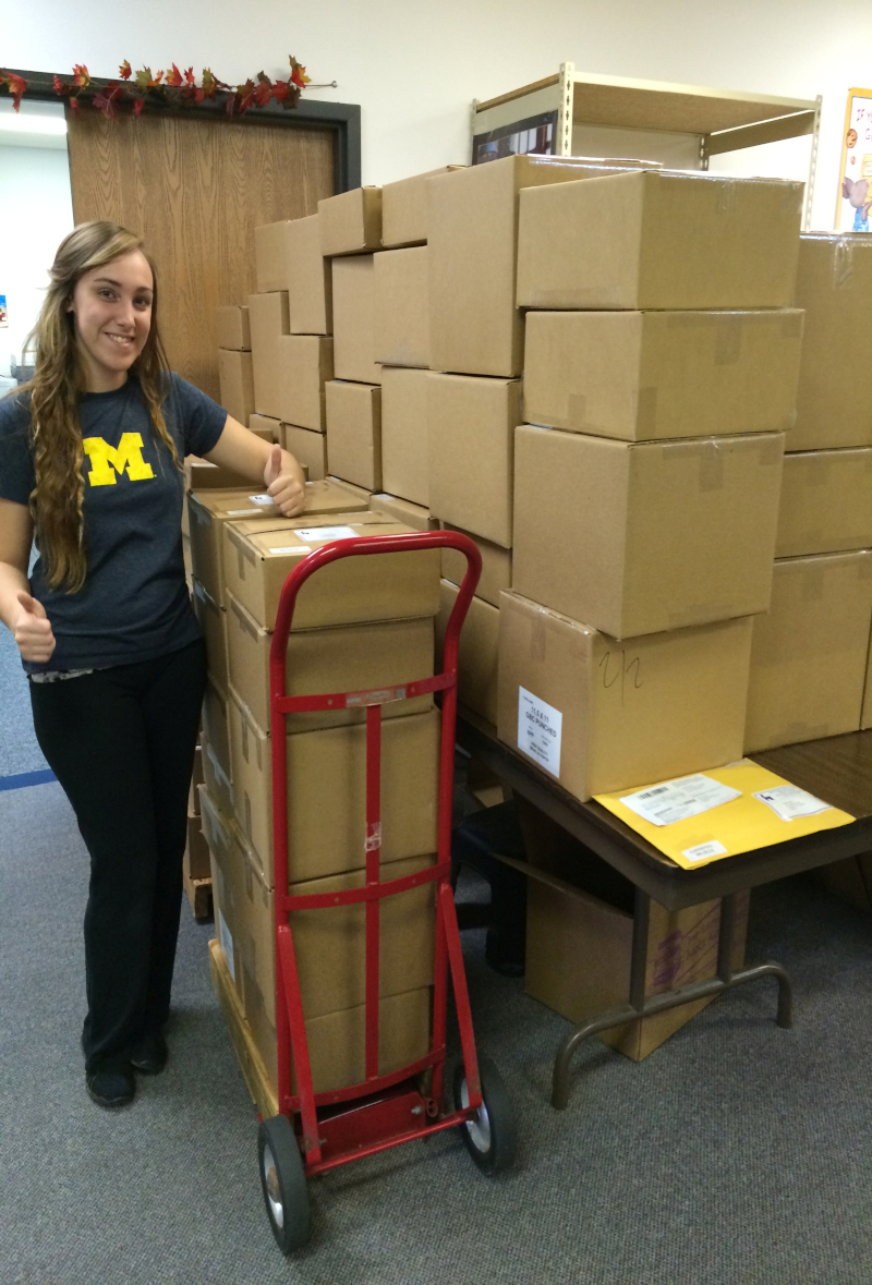 Seedlings' former employee, Katelynn, giving the camera two thumbs up while standing next to a table full of boxes of braille books.