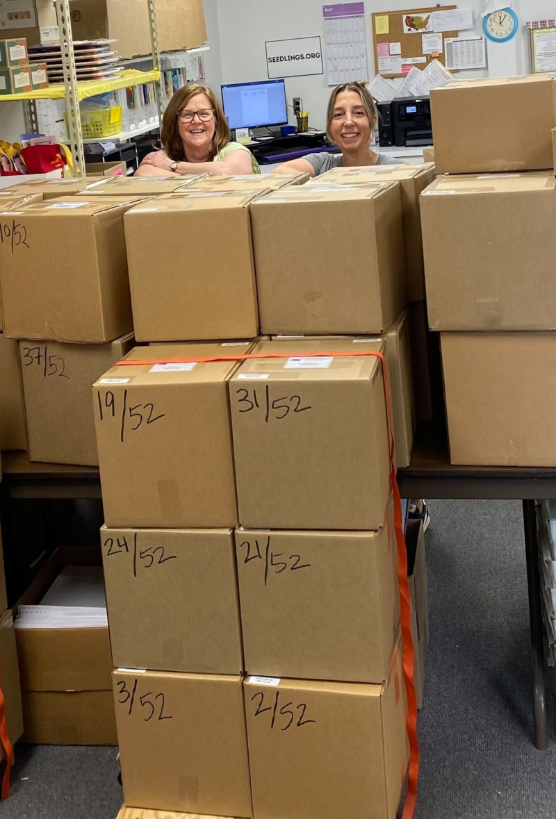 Seedlings' employees, Lori and Mary, smiling faces peak out behind a large 52 box shipment of braille books.