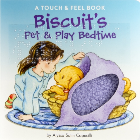 A young child, wearing pajamas, sits with Biscuit, the puppy. Biscuit sits in a basket and the child is covering him with a blanket.